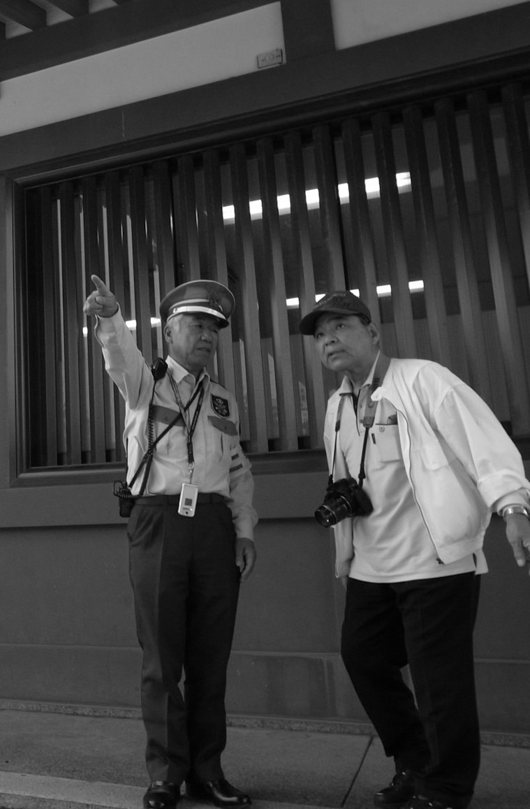 An official-looking gentleman in a peak cap and wearing a lanyard and walkie-talkie, directs a second gentleman who appears to be a tourist (he has a big camera around his neck and is casually dressed) by pointing in the desired direction.