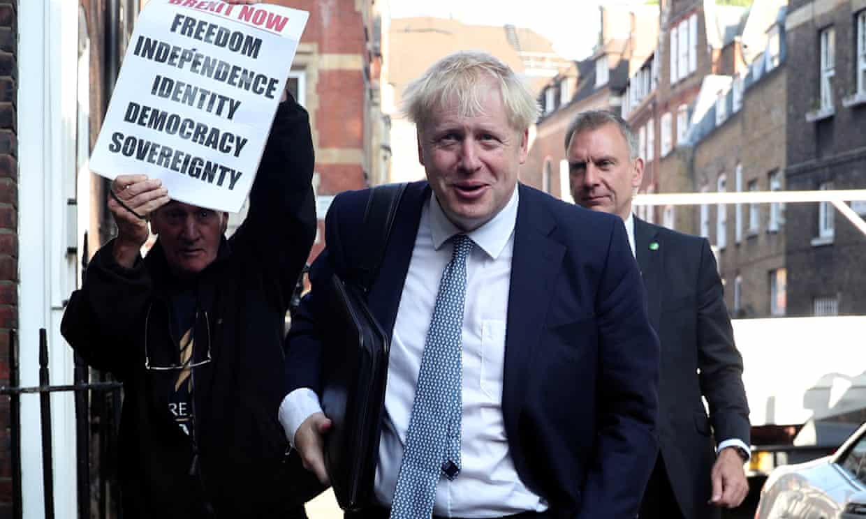 Boris Johnson in a London street. Behind him a man holds a poster that reads Brexit Now - freedom, independence, identity, democracy, soverignty