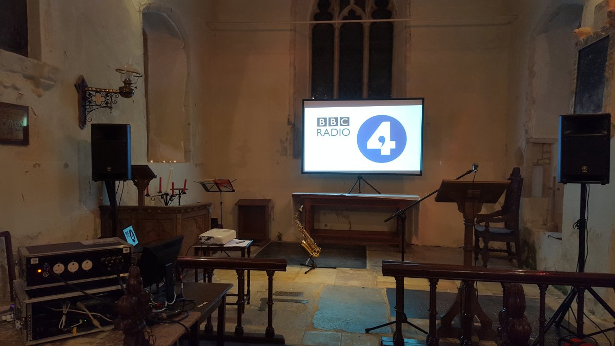 The altar area of a small church. On either side, there is a speaker on a stand; there's a microphone on a stand by the lecturn and a good deal of audio equipment to the left hand side. In front of us is the screen on which is being projected the Radio 4 logo. There are no people in the picture - something's about to happen!