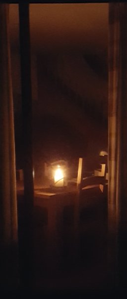 A candle is on a dining table, seen from outside the house looking in on a dark night