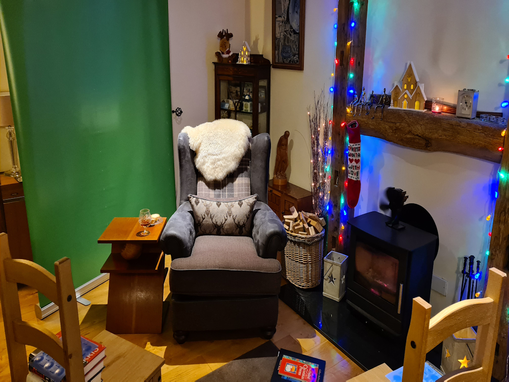 An armchair next to a log burner, surrounded by Christmas decorations. To the other side of the chair is a table on which we see a glass of brandy and a couple of mince pies. Behind the chair is a green screen