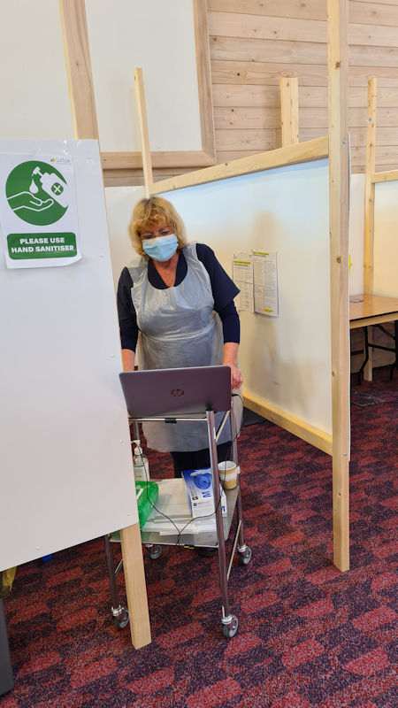 A nurse in the entrance to a small booth, looking at her laptop on top of a small trolley. The patient she's talking to is inside the booth and hidden