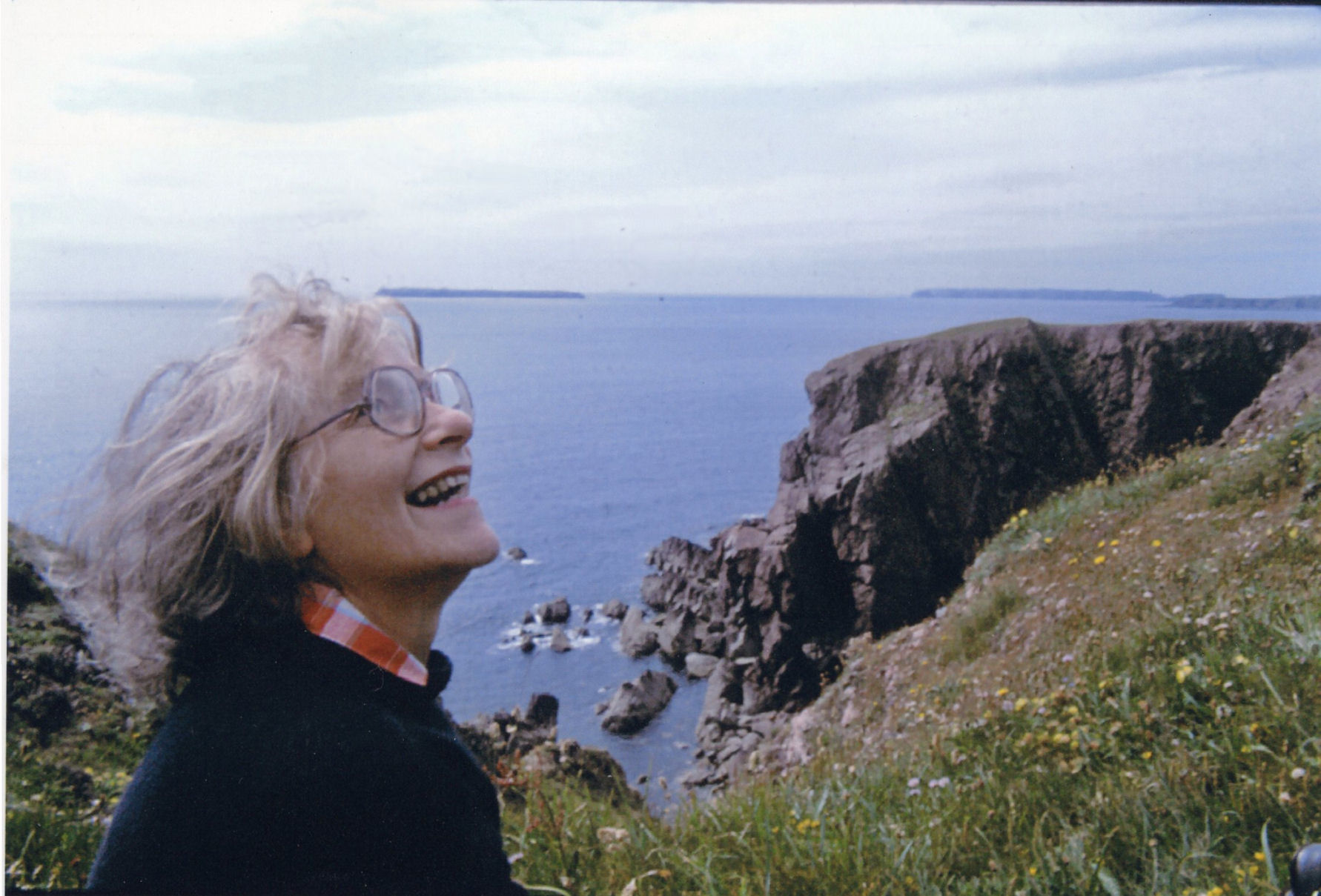 A middle aged woman is smiling looking up at something in the sky, against a backdrop of a rocky coastline with islands in the background